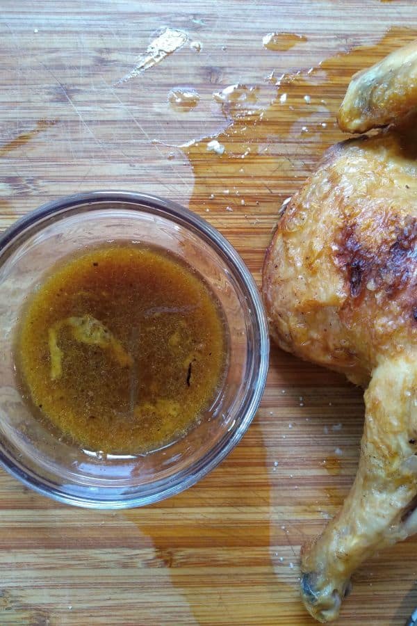 A small glass bowl of au jus on a cutting board next to a piece of chicken.