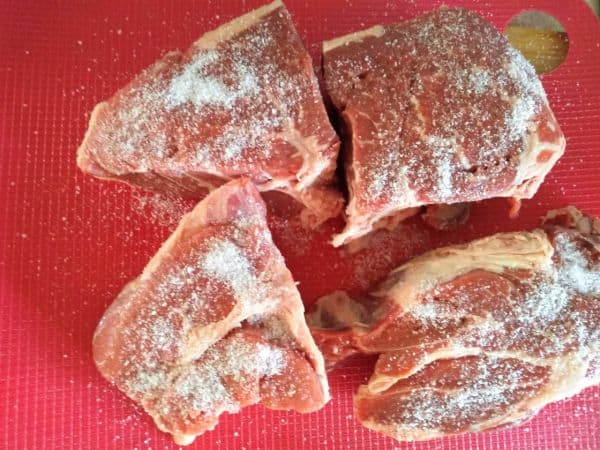 A boneless chuck roast is cut into 4 pieces and covered in kosher salt. The pieces are on top of a red cutting board.