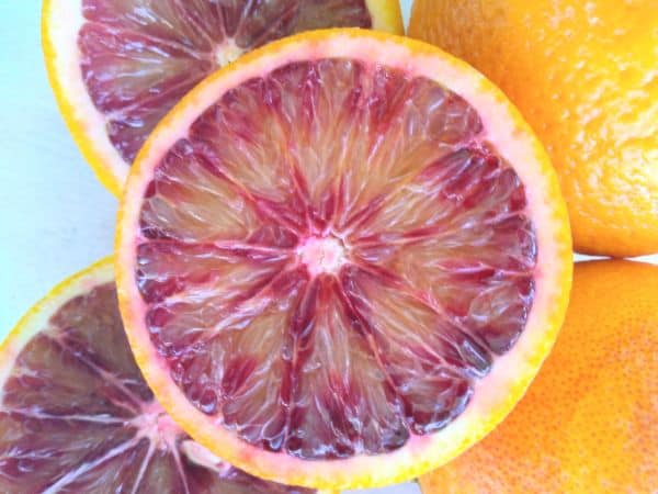 Blood oranges are cut in half to show the inside while sitting on top of whole blood oranges.