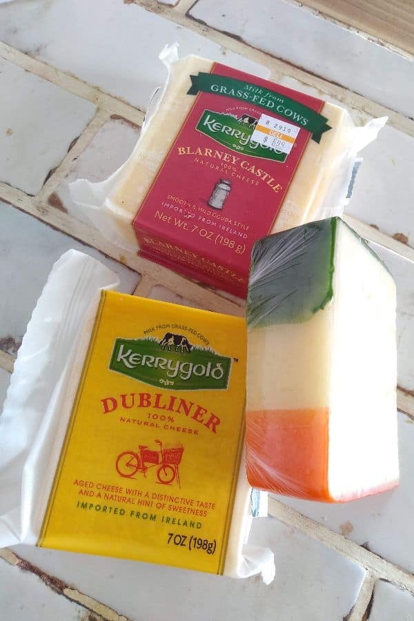 Kerrygold blarney castle cheese, Dubliner cheese, and a cheese with wax that represents the color of Ireland (green, white, and orange) sitting on a white tile countertop.