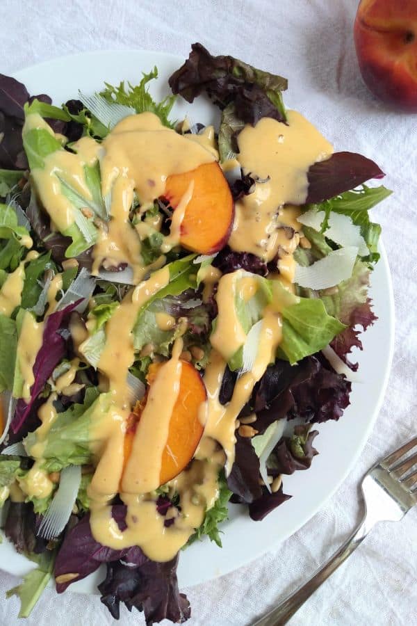 Peach tahini salad dressing on a salad with fresh peaches and spring mix on a white plate. A fork is next to the plate and the plate is on a white towel.