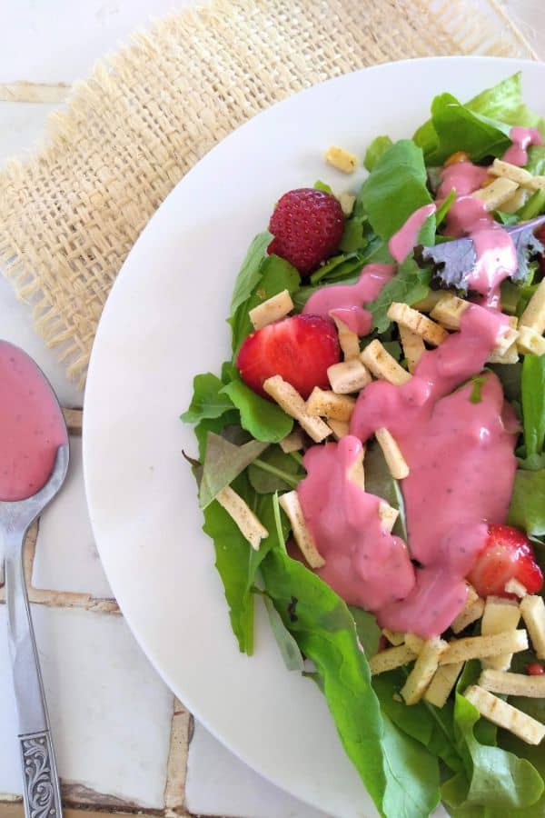 A mixed greens salad with fresh strawberries and wonton strips topped with pink strawberry tahini dressing. The salad is one a white plate on top of a piece of burlap on a white tile countertop.