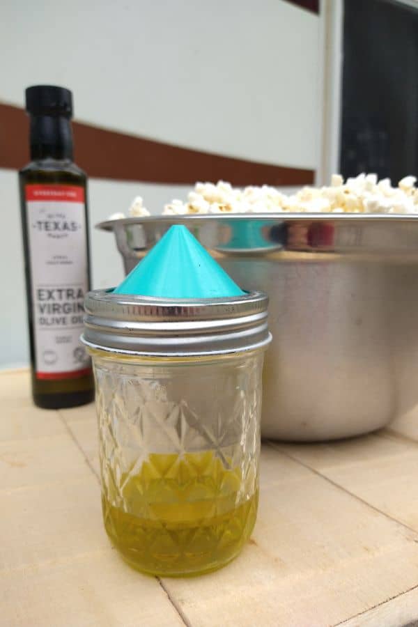 A mason jar with olive oil with the Mini ergo spout lid attached sitting on a wood board with a bottle of Texas Extra Virgin olive oil and a bowl of popcorn in the background