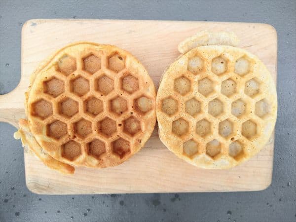 Two waffles sitting on a wood cutting board. The waffle on the left is more brown than the waffle on the right.