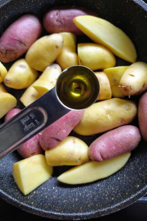 Sliced fingerling potatoes in a frying pan with a teaspoon measuring of olive oil over top ready to be added to the pan.