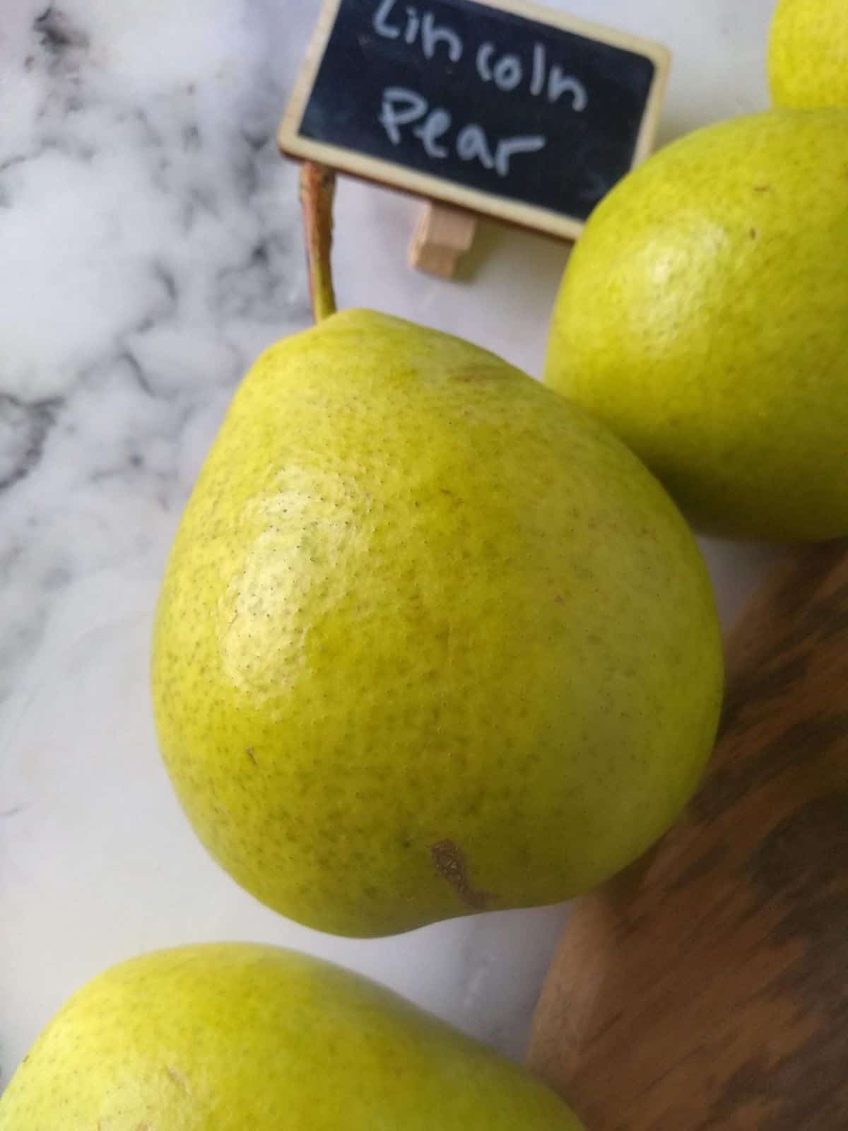 Lincoln pears sitting on a white surface next to a cutting board with a  small chalkboard sign that say what variety they are.