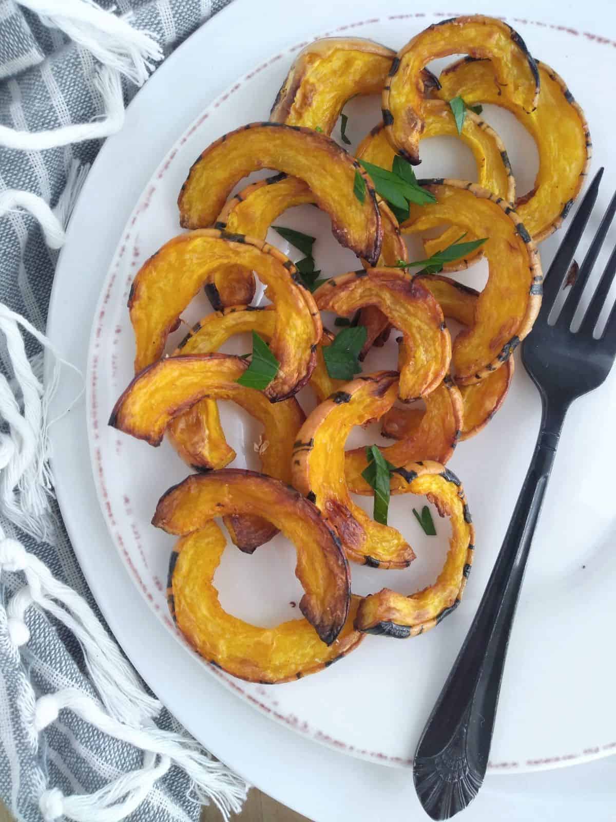 A plate of Roasted Delicata squash pieces with the skin on on top of a larger plate on top of a gray and white striped towel. A black fork is next to the squash.