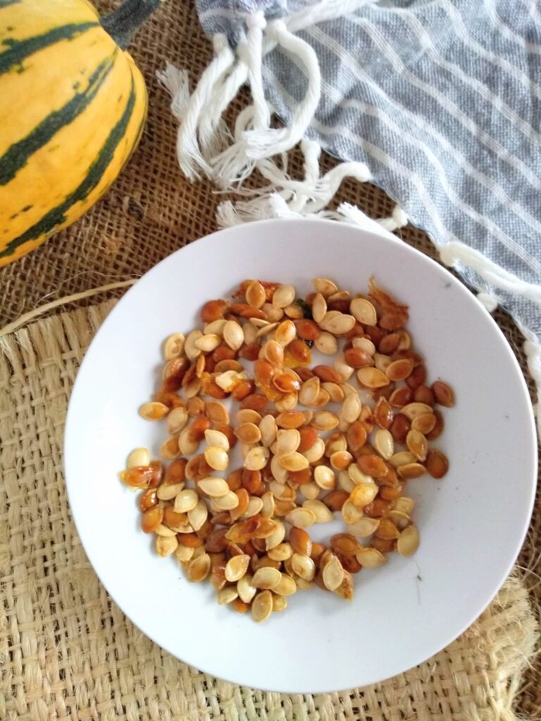 A white plate with roasted Delicata squash seeds sits on a piece of light brown burlap on top of darker brown burlap with a gray striped towel to the right and a yellow Delicata squash in the upper left corner.