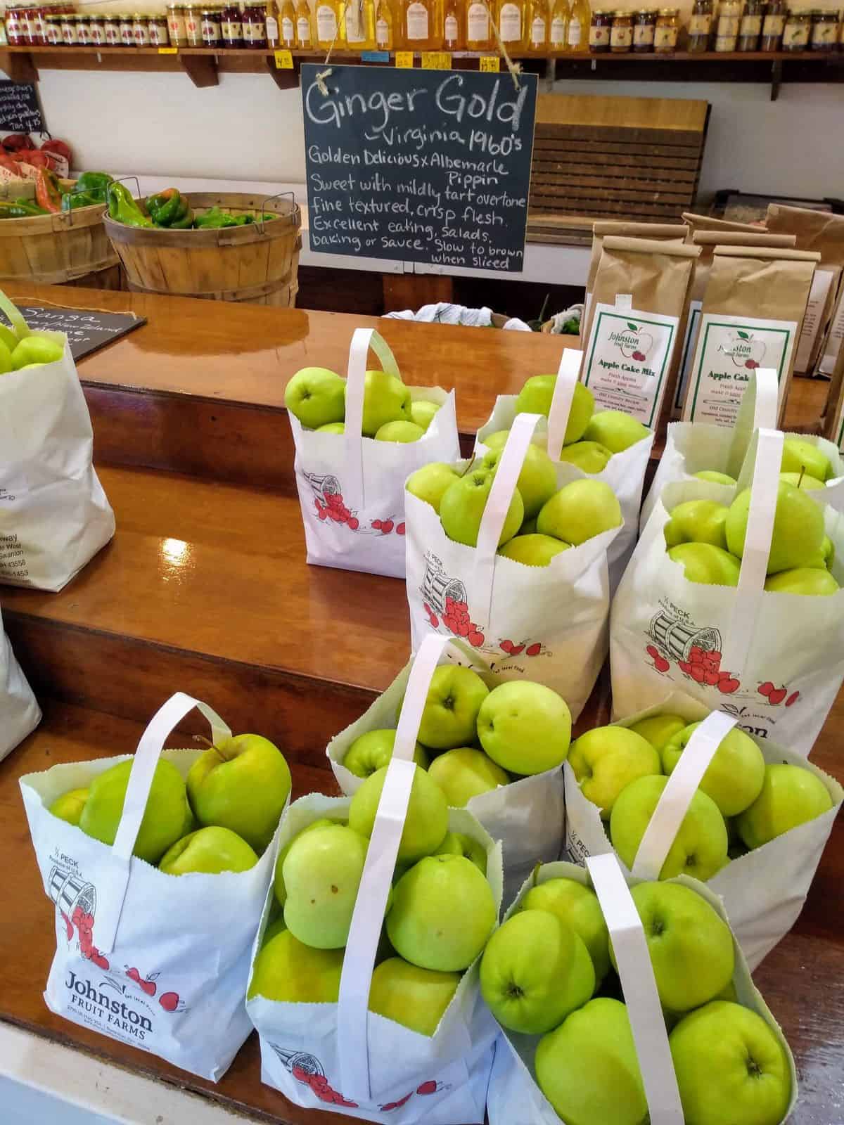 Bags of Ginger gold apples sit on a layered shelves at a farm market. The sign describes them as a cross between Golden Delicious and Albermale Pippin. Sweet with mildly tart overtone. Fine textured, crisp flesh. Excellent eating, salads, baking or sauce. Slow to brown when sliced.