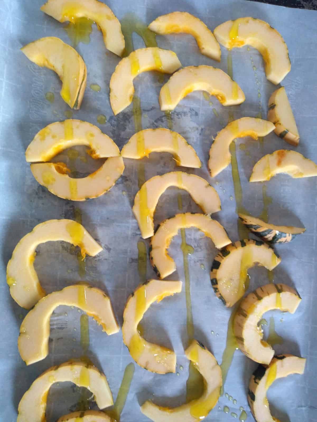 Half moon shaped Delicata squash on top of a parchment paper with olive oil drizzled on top.