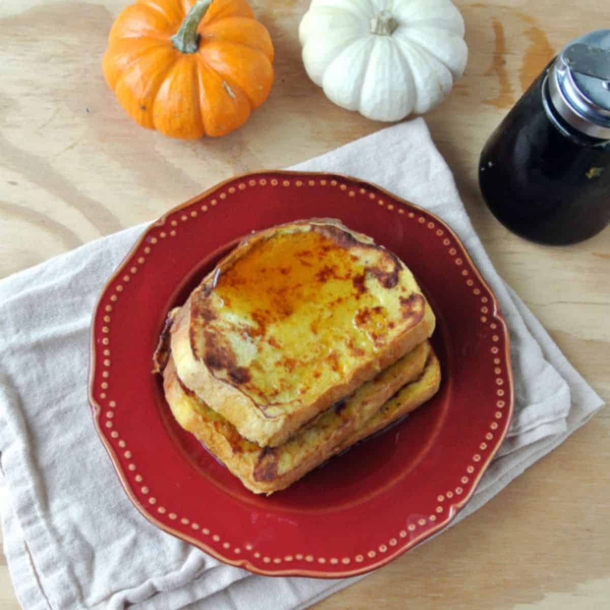 Three slices of pumpkin french toast on a red plate on top of a white towel. Nex to the plate is a container of maple syrup, 1 orange mini pumpkin, and 1 white mini pumpkin