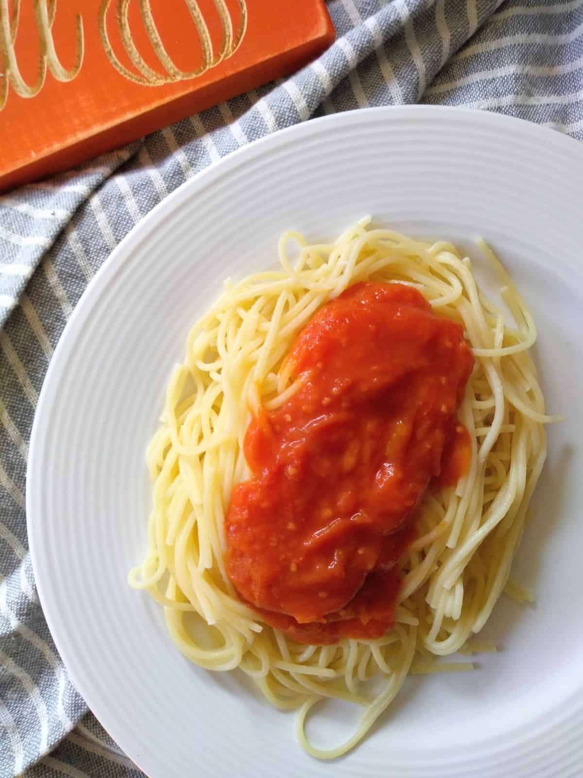 A white plate with spaghetti on it topped with a tomato sauce made with tomatoes and some pumpkin puree. The plate is on a gray and white striped towel with a wood sign at the top left corner that is orange with a pumpkin on it. 