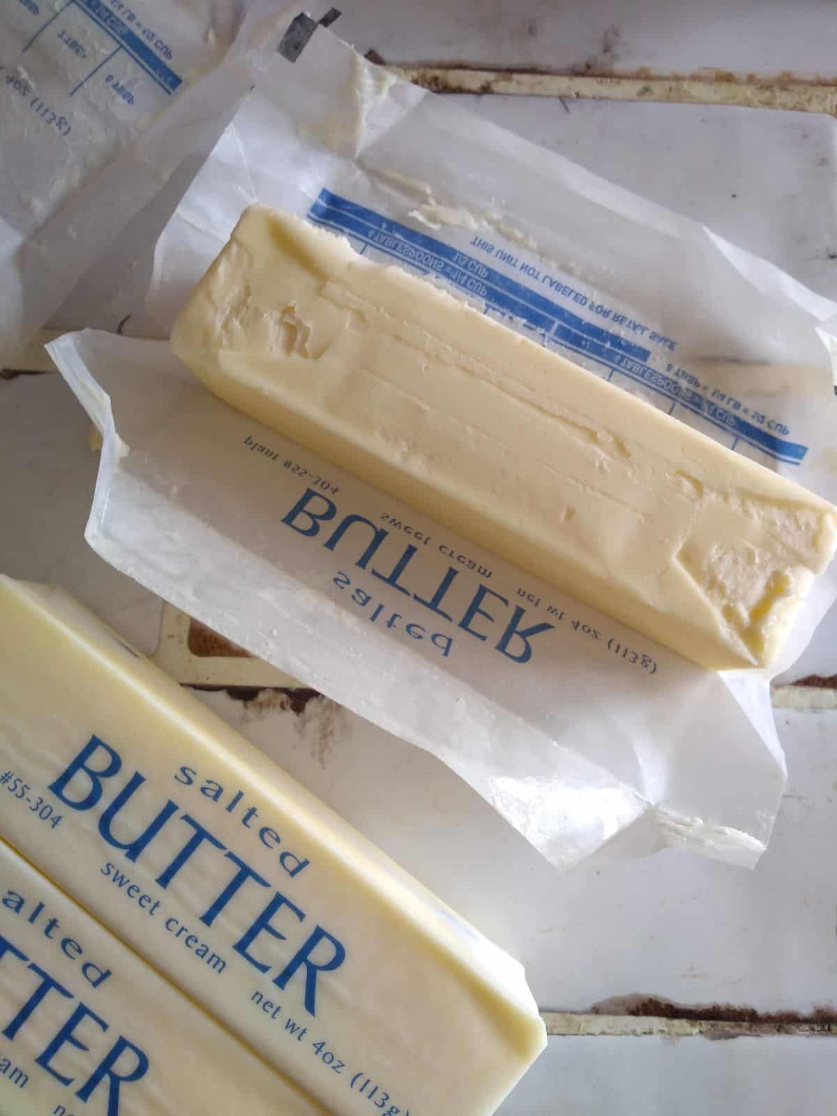 On a white title counter top is 3 sticks of butter. The butter at the top is opened and sitting on it's wrapper while the other 2 sticks are still in their wrappers.