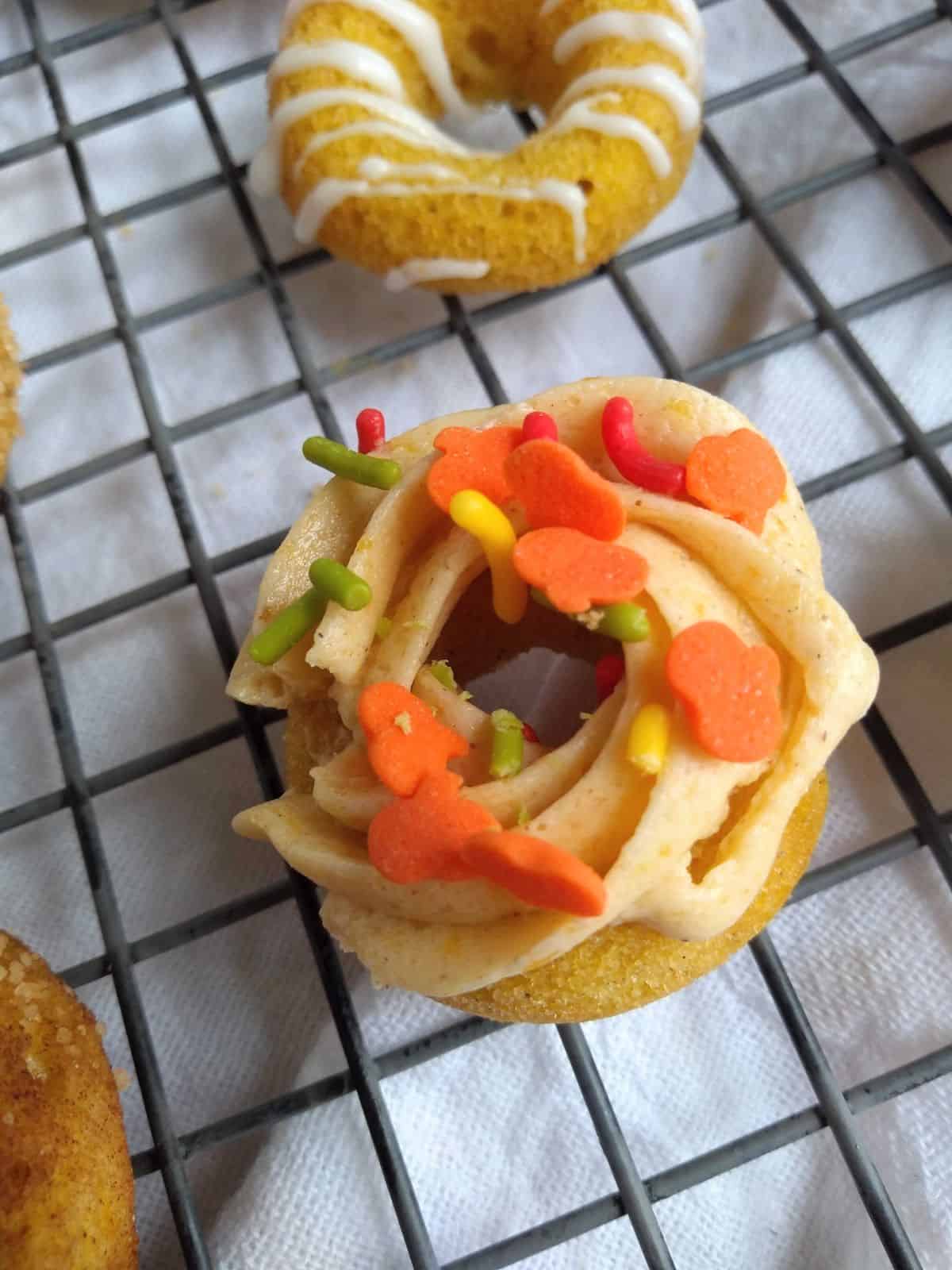 Mini pumpkin donuts on a wire rack over a white towel. The donut in the middle has pumpkin buttercream frosting and pumpkin & fall colored sprinkles on top.