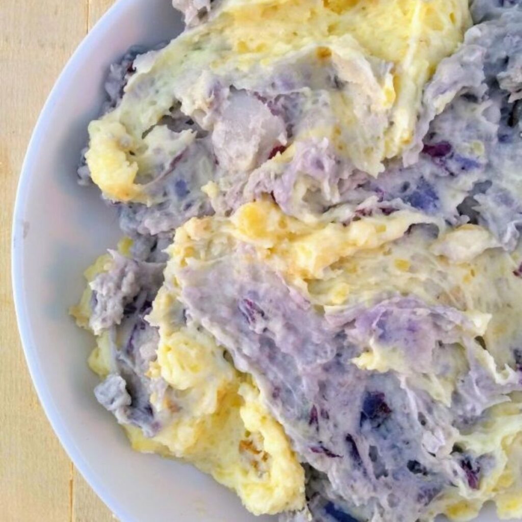 Gold mashed potatoes and purple mashed potatoes swirled together in a white bowl on top of a wood board.