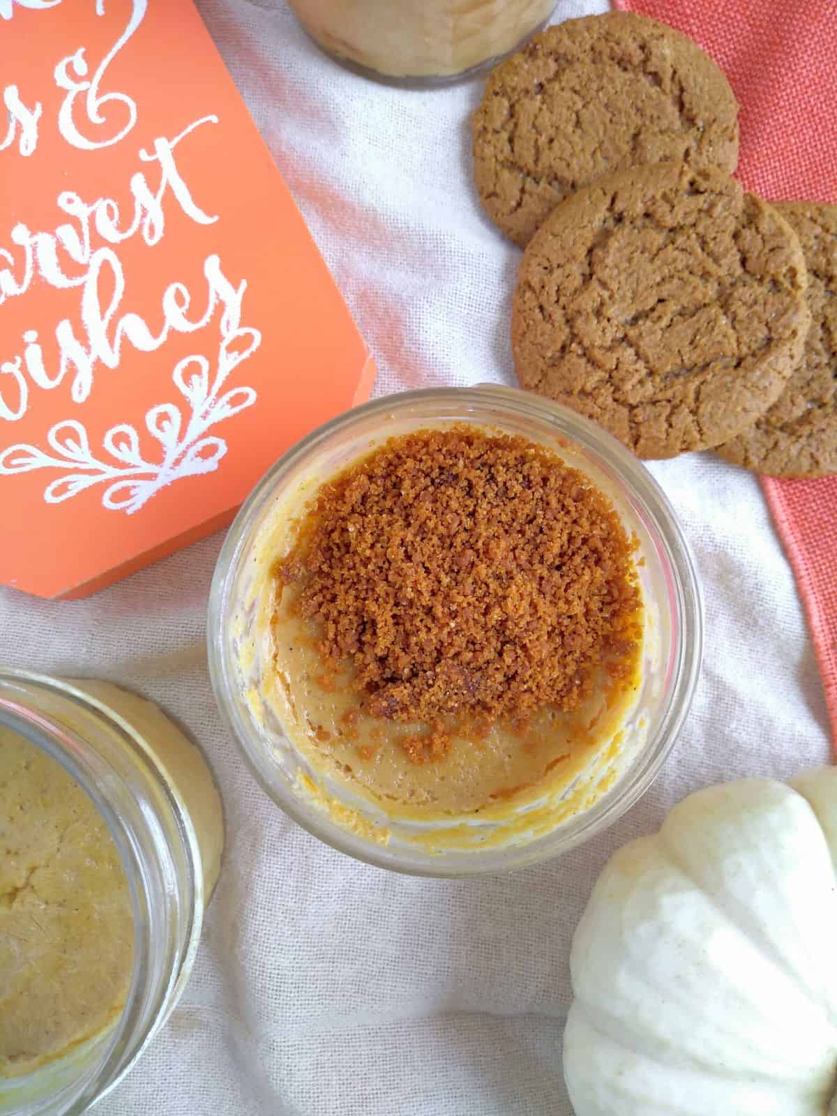 A jar filling with sous vide pumpkin filling topped with a gingersnap crumble. Ginger snap cookies, a orange colored sign with handwriting on it, a jar of pumpkin filling with no crumble, and a mini white pumpkin all around the crumble topped jar.