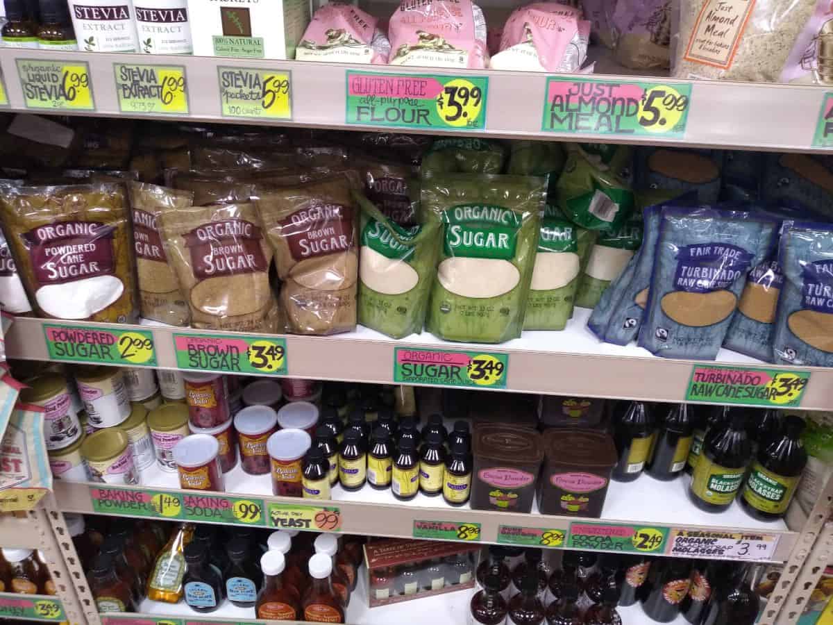 A picture of a section of the bakery section at Trader Joe's store. The items pictured on the shelf include powdered sugar, brown sugar, cane sugar, turbinado sugar, almond meal, alternative sweeteners, vanilla extra, and more.