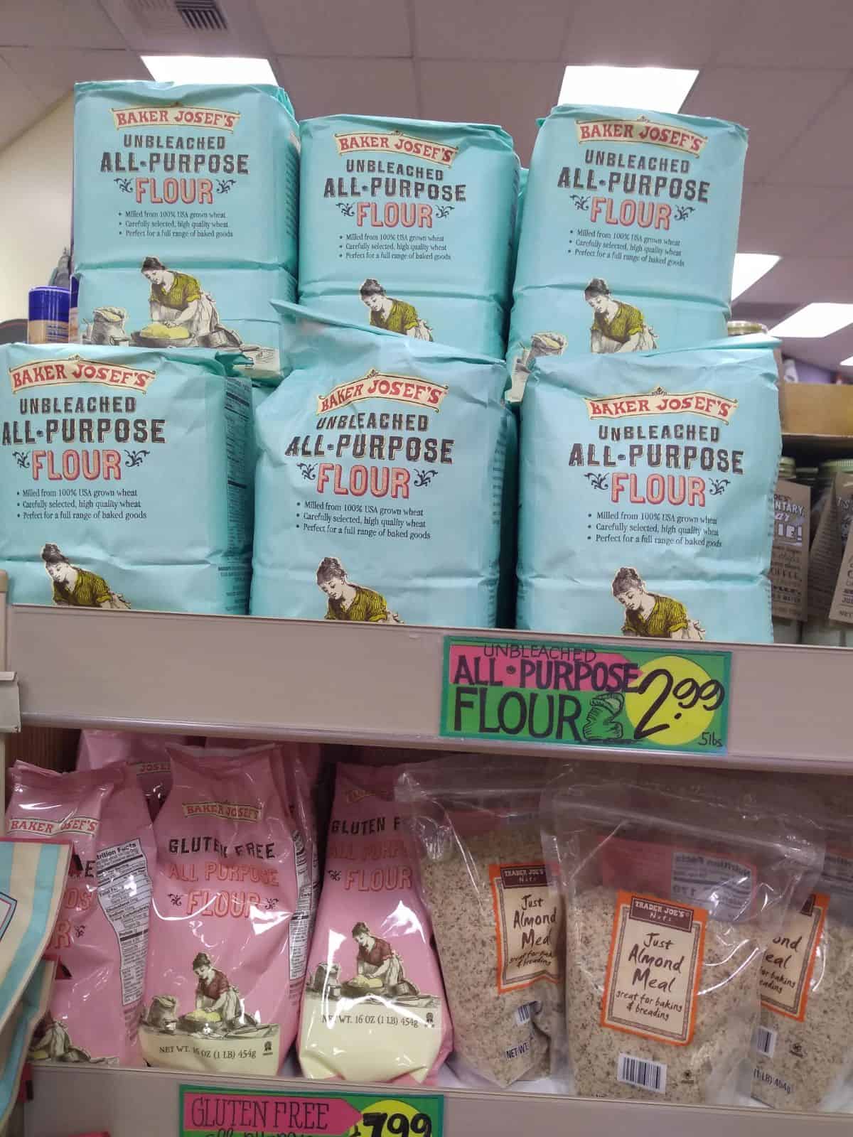 Bags of Baker Josef's Unbleached All Purpose Flour sit on the shelf above  bags of gluten free flour and Just Almond meal.