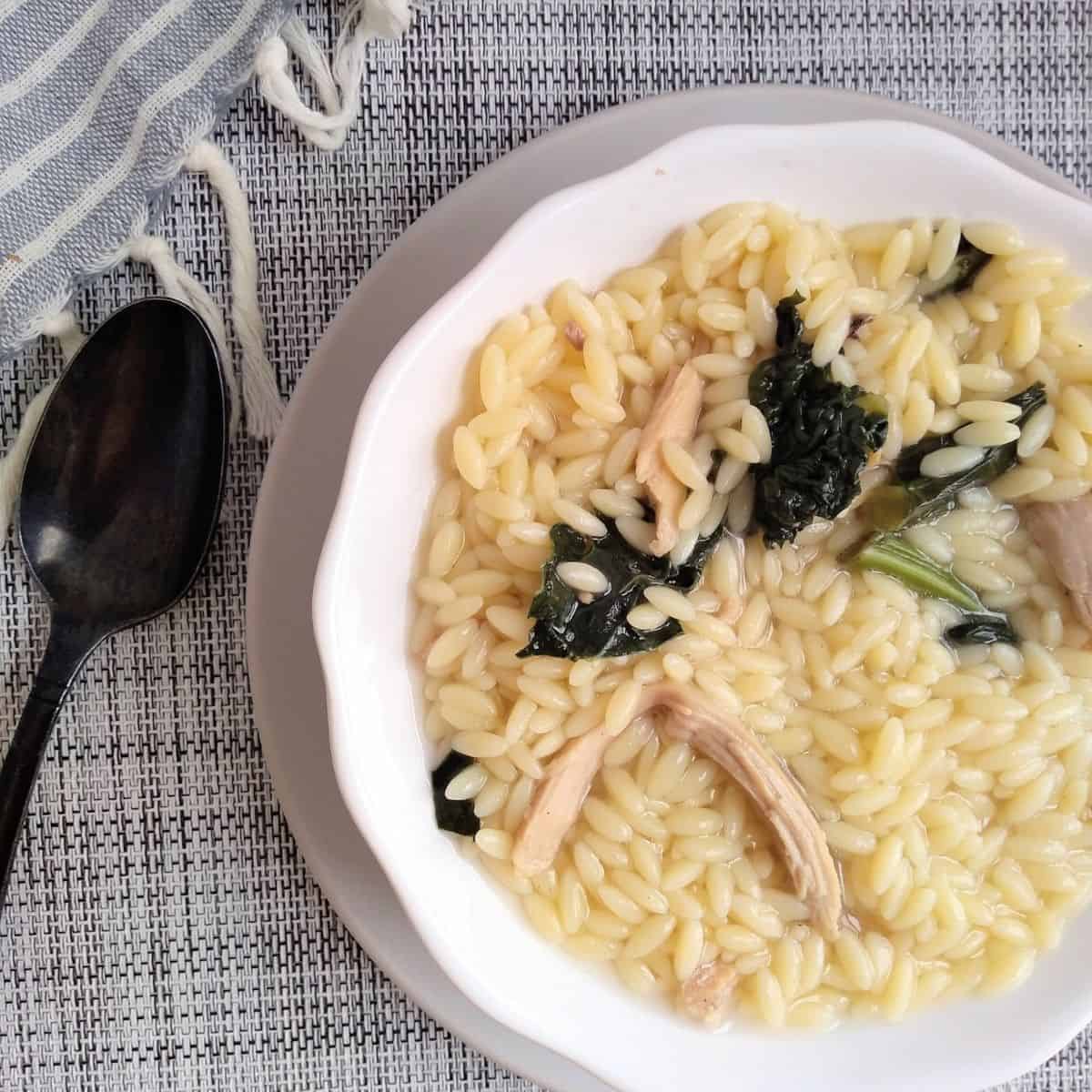 A white bowl on a gray plate filled with leftover turkey, orzo and spinach in a turkey broth. Next to the bowl is a black spoon. 