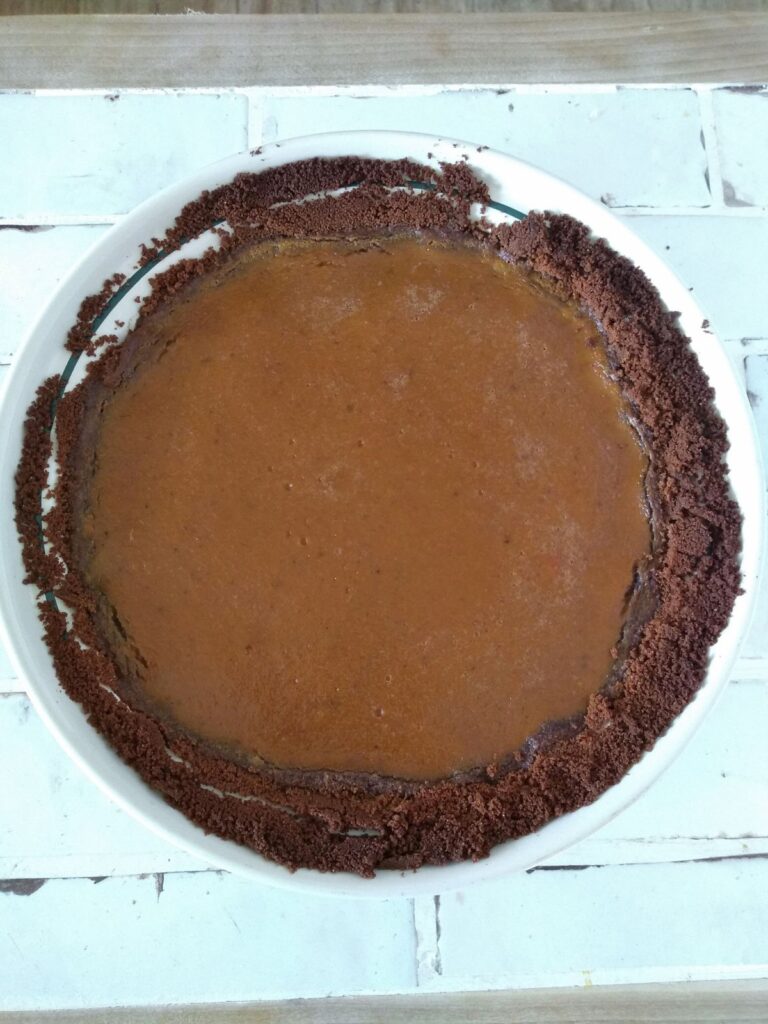 An overview of a pumpkin pie with a gingersnap crust. The pie is sitting on a white tile countertop. The crust has separated from the pie in the top left corner.