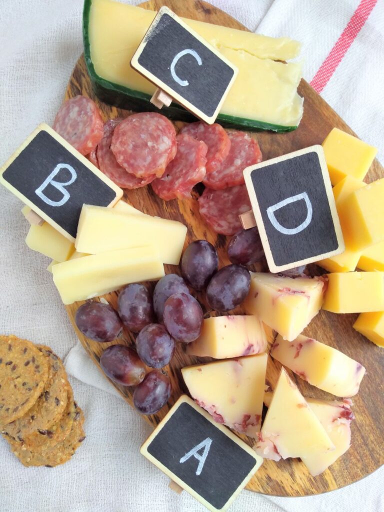 A cheese board with 4 different types of cheeses in different shapes with small chalk board tags with letters A,B,C, and D on them. Grapes and salami are on the board as well with crackers next to it.