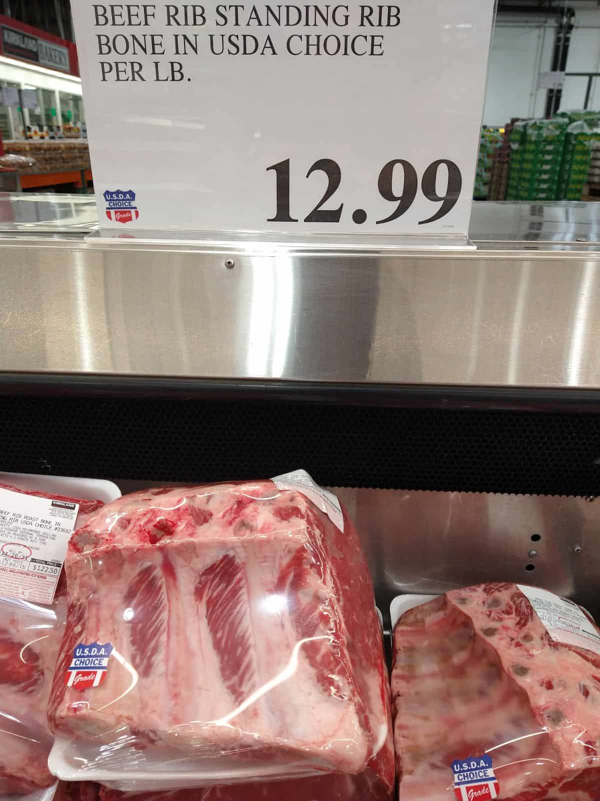 A sign reads "Beef Rib Standing Rib Bone In USDA Choice Per lb 12.99. Piles of roasts underneath the sign in the cooler.