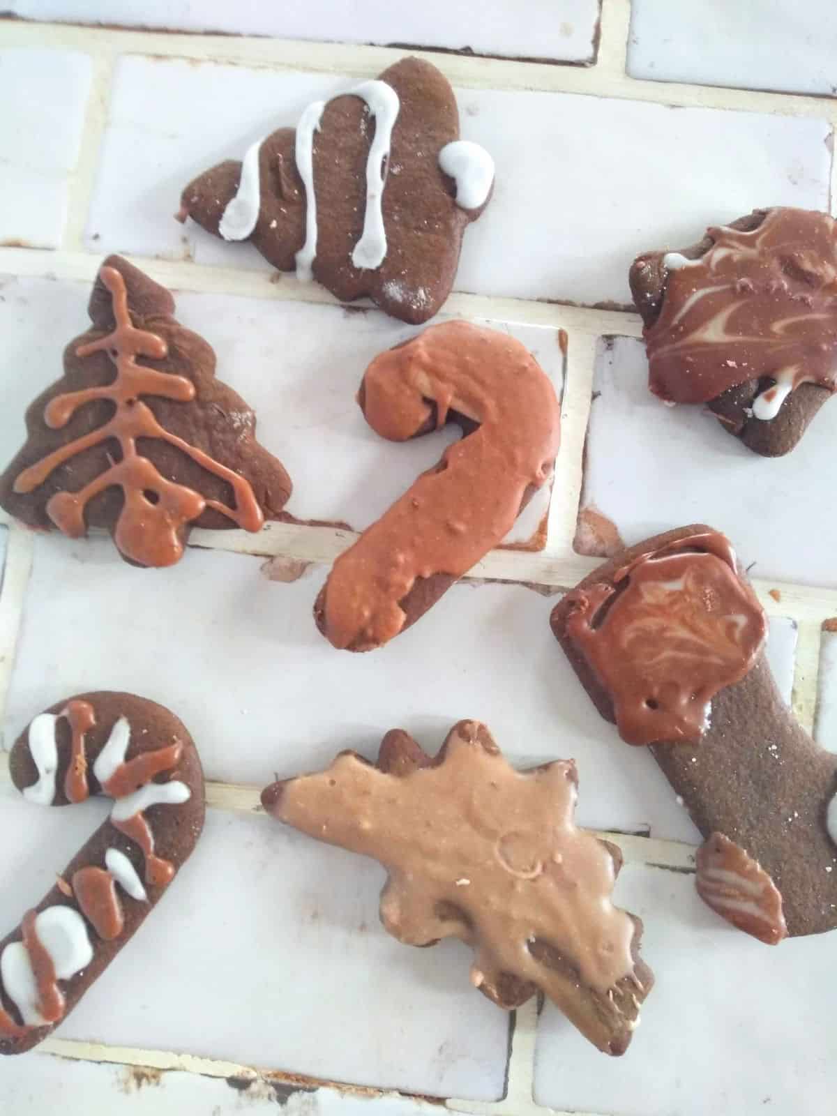 Small cut out ginger cookies in shapes lie trees, candy canes, stockings, stars, bells, and a dinosaur. The cookies are frosted with different colored chocolate frostings and are sitting on a white tile countertop.