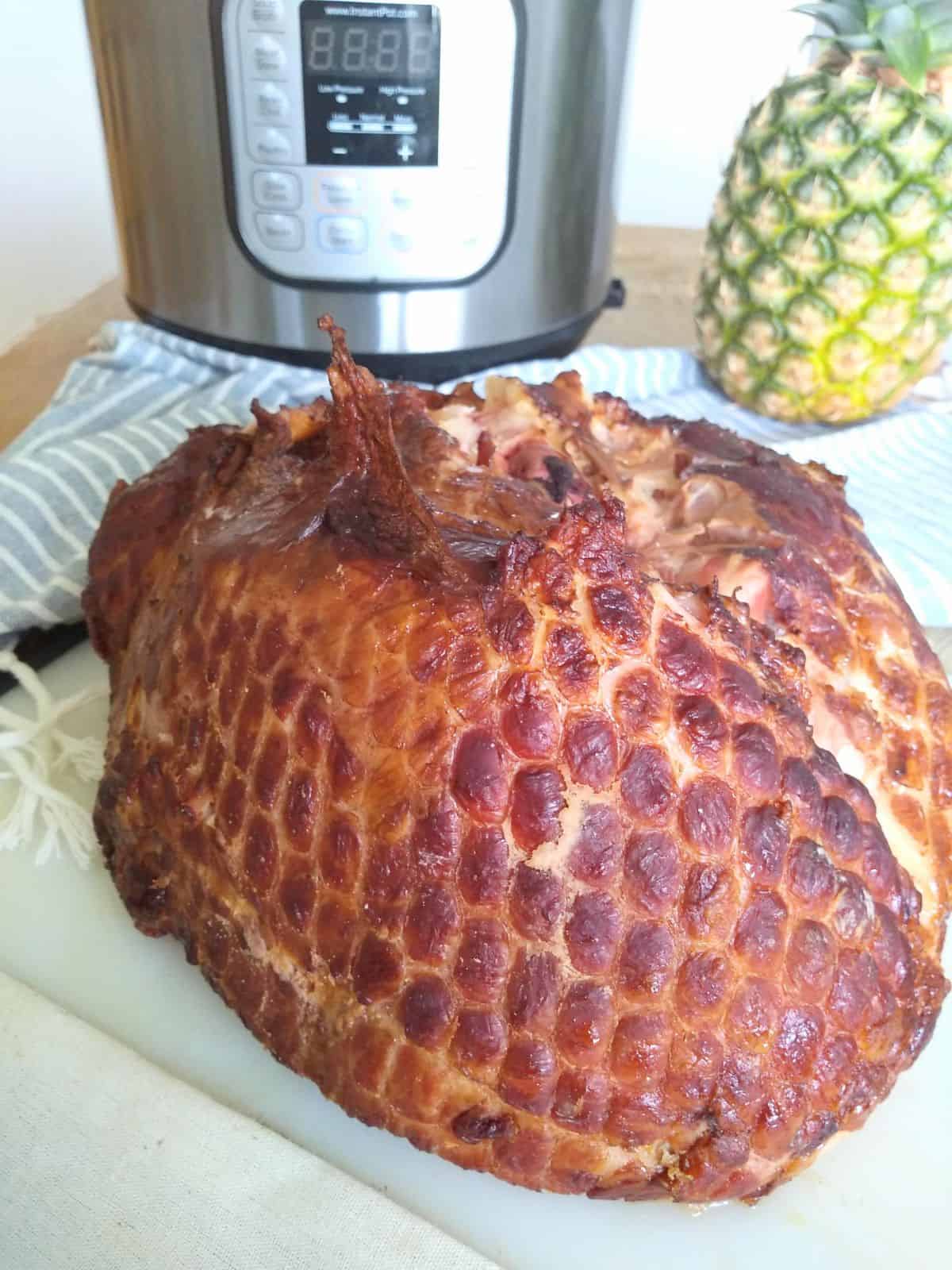 A cooked ham sitting on plastic cutting board with an Instant Pot, a whole pineapple, and a gray ahd white stripped towel in the background.