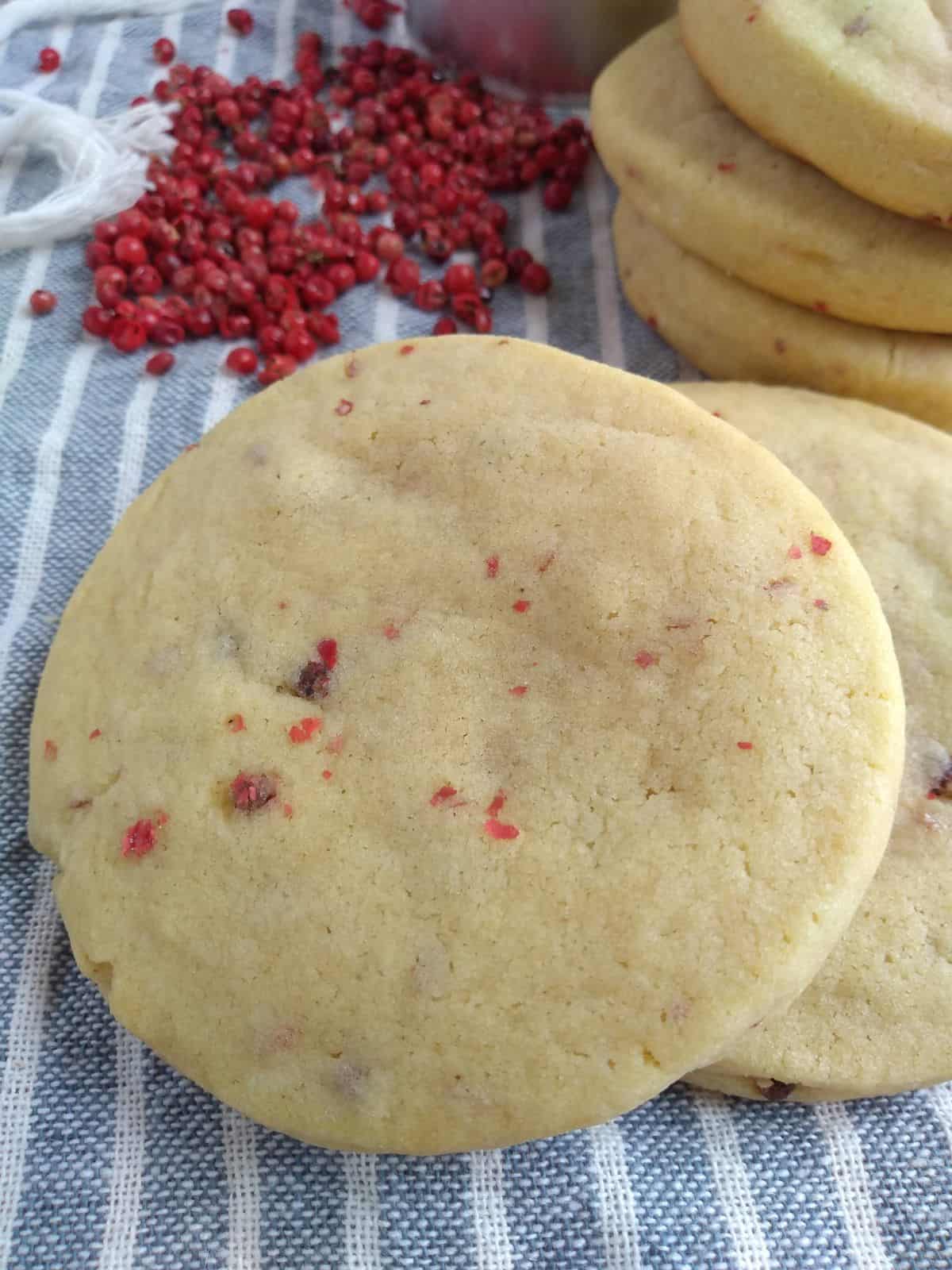 A sugar cookie filled with pink peppercorns on top of other cookies with a stack of the same cookies in the background. The cookies are on top of a gray striped towel with pink peppercorns in the top left corner.