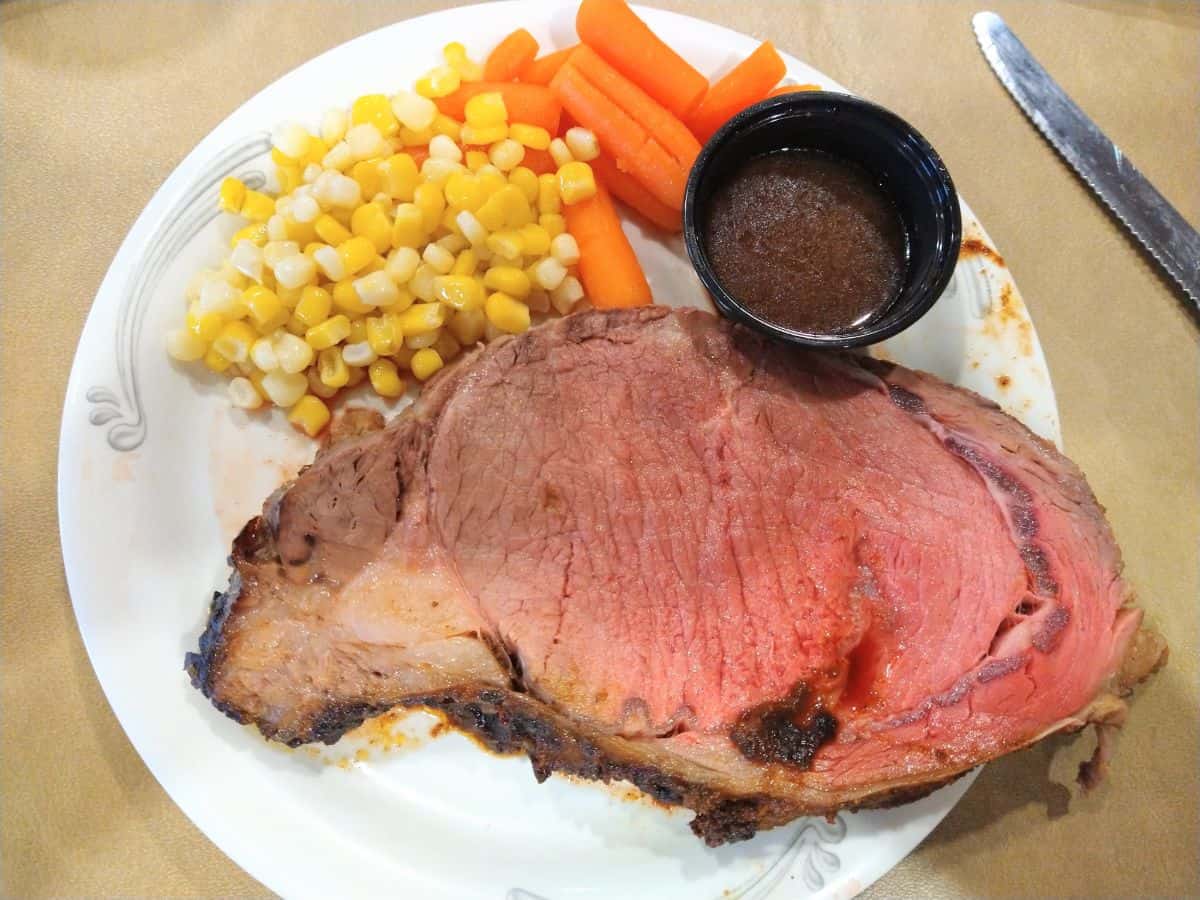 A piece of prime rib on a paper plate with corn, carrots, and au jus in a small black container on the plate as well.