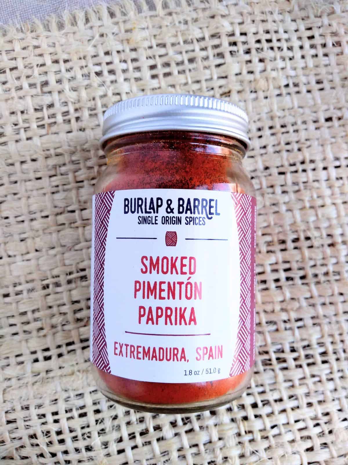 A glass jar of Burlap & Barrel Single Origin Spices Smoked Pimenton Paprika from Extremadura, Spain. The jar is on top of a piece of light brown burlap.