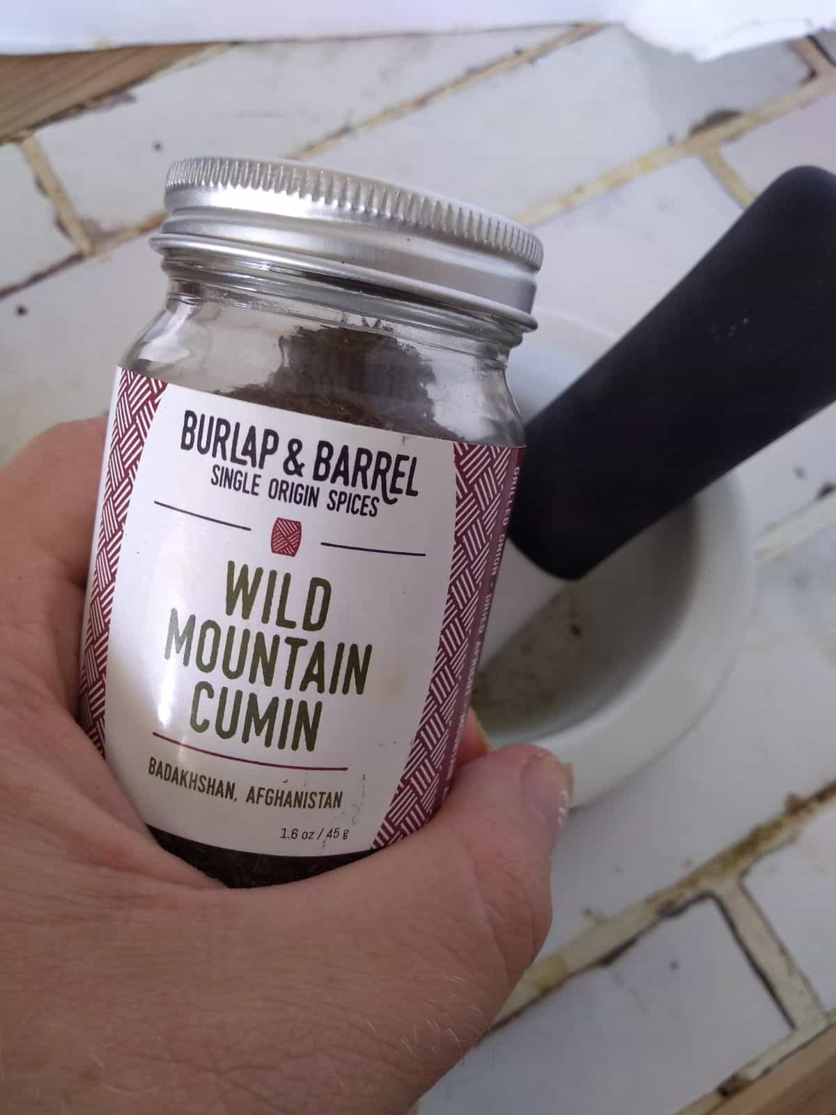 A glass jar of Burlap & Barrel Single Origin Spices Wild Mountain Cumin from Badakhshan, Afghanistan. The jar is being held over top of a mortal and pestle. 