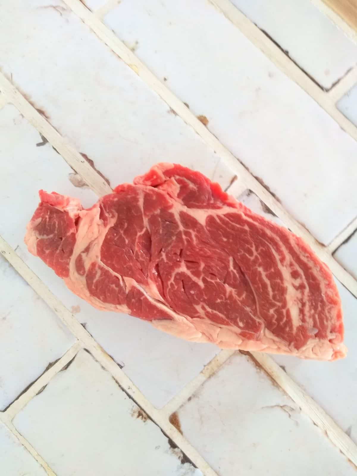 A raw center eye chuck eye steak sitting on white tile countertop.
