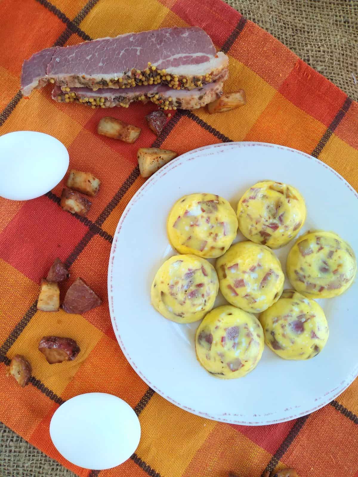 A overhead view of corned beef hash egg bites on a white plate with brown trim on top of a orange plaid towel with an egg and corned beef and potatoes surrounding the plate.