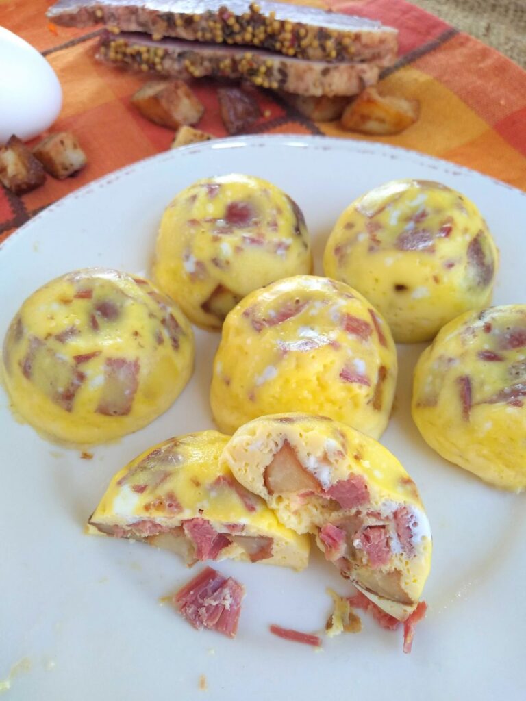 Corned beef hash egg bites on a white plate with brown trim. One egg bite is cut open showing the potato and corned beef inside. Corned beef, potatoes and an egg are shown at the top above the plate.