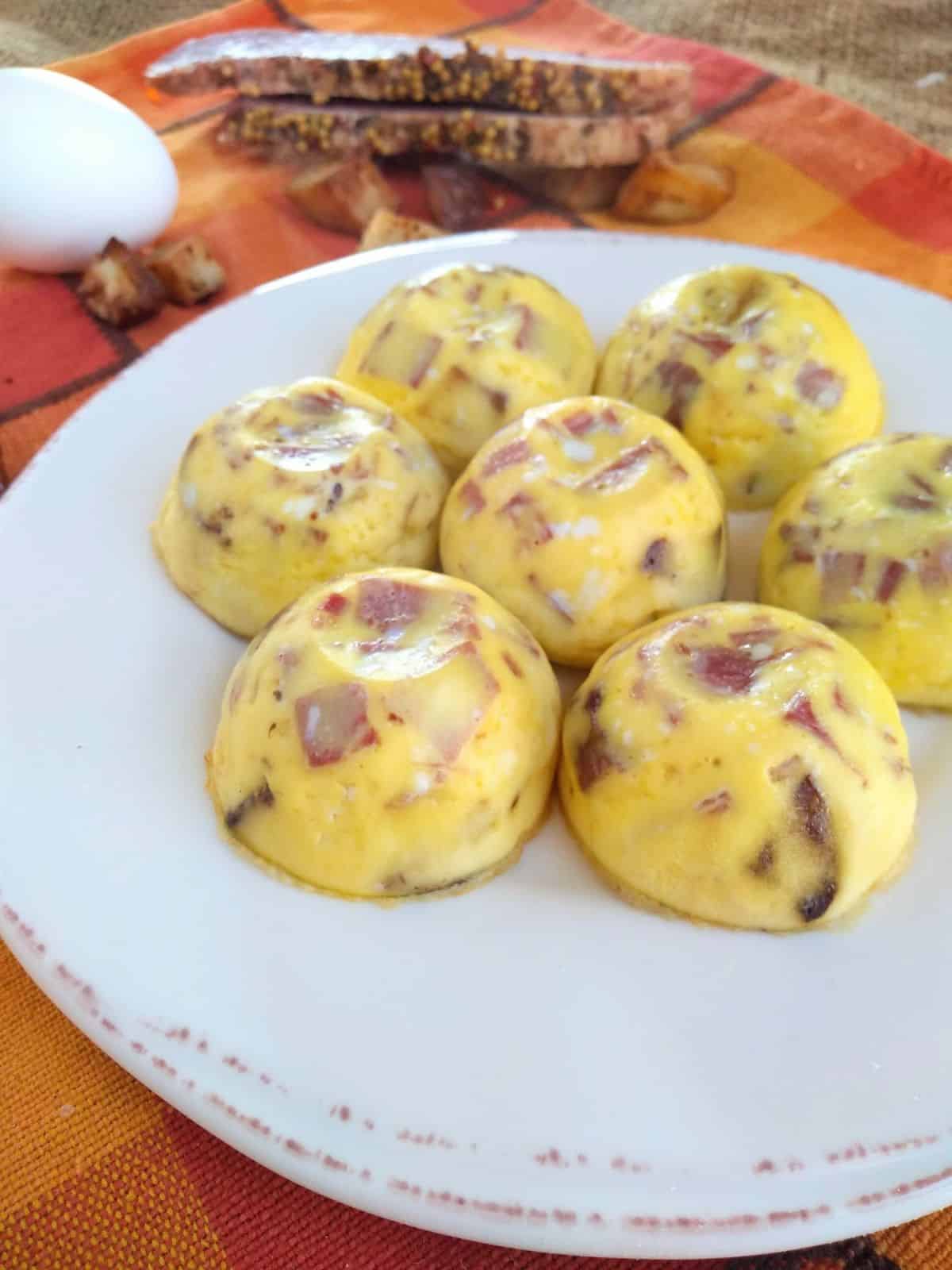 Corned beef hash egg bites on a white plate with brown trim on top of a orange plaid towel with an egg and corned beef and potatoes at the top.