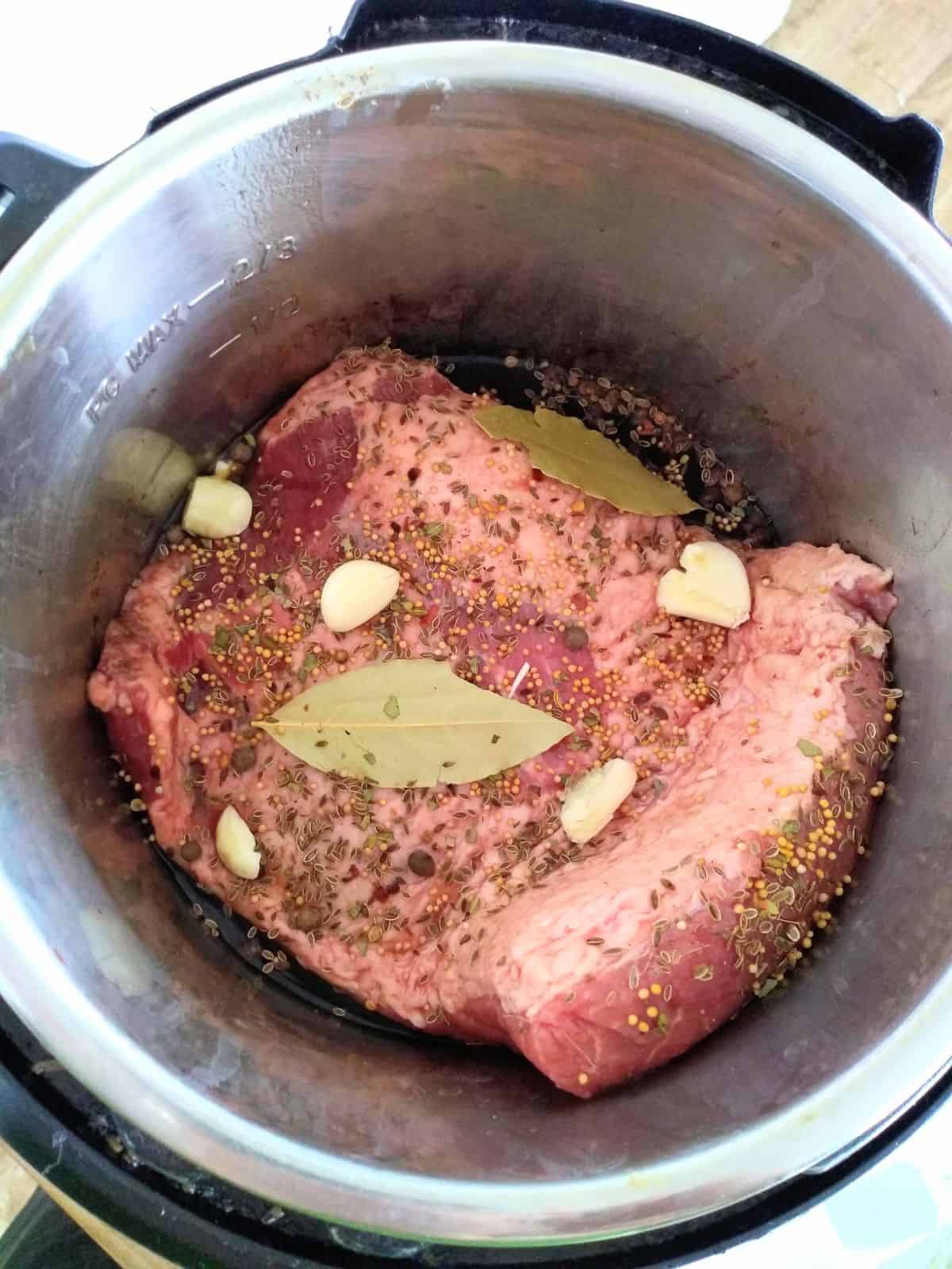 Corned beef in the Instant Pot in liquid with garlic cloves, bay leaves, and spices on top.