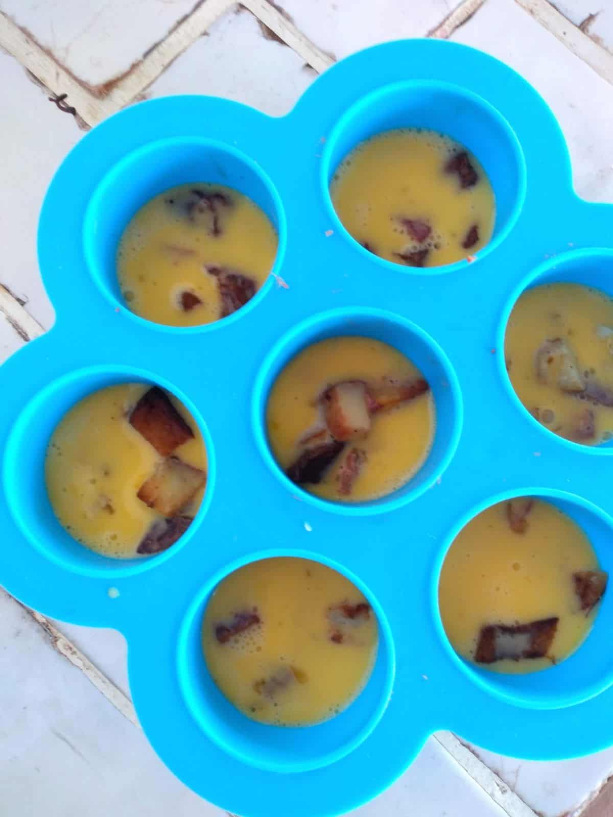 A blue silicone egg bite mold filled with scrambled raw eggs with potatoes and corned beef inside. The mold is sitting on a white title countertop.