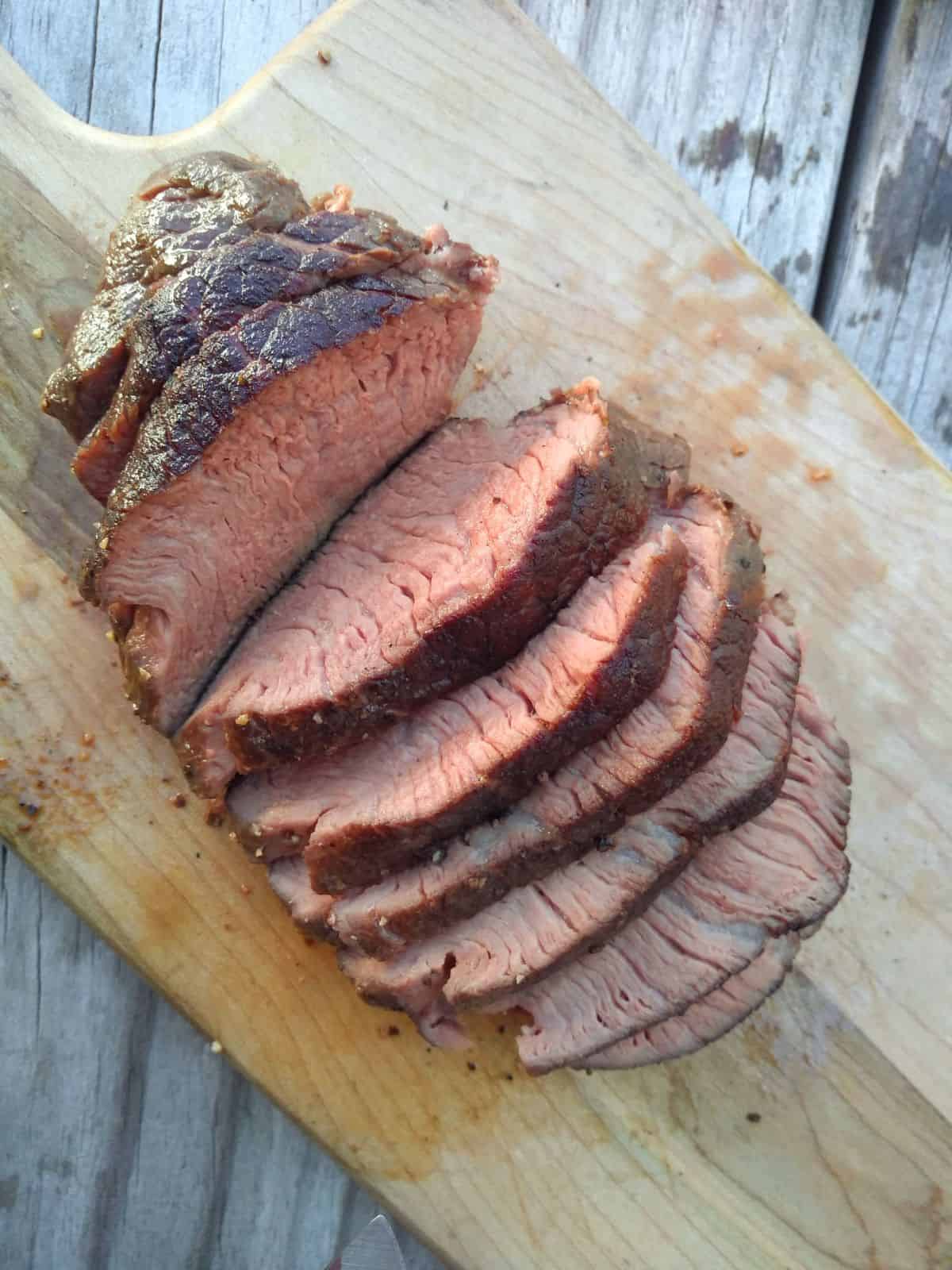 A browned Sierra steak that is perfectly pink from edge to edge is arranged in slices on a wood cutting board.