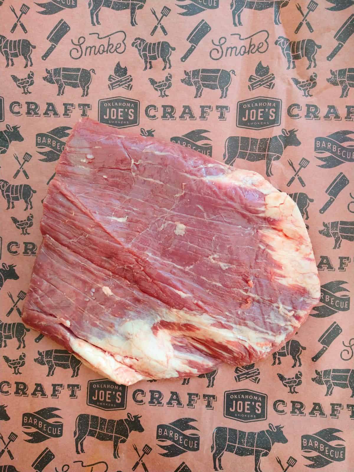 A raw flank steak sitting on a piece of reddish colored butcher paper with grilling images on it. 