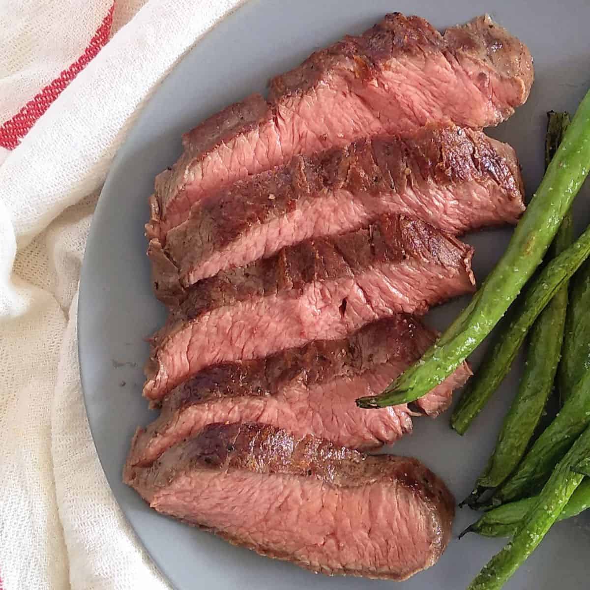 A flat iron steak cut into pieces on a blue plate with green beans. The plate is on a white towel with a red stripe