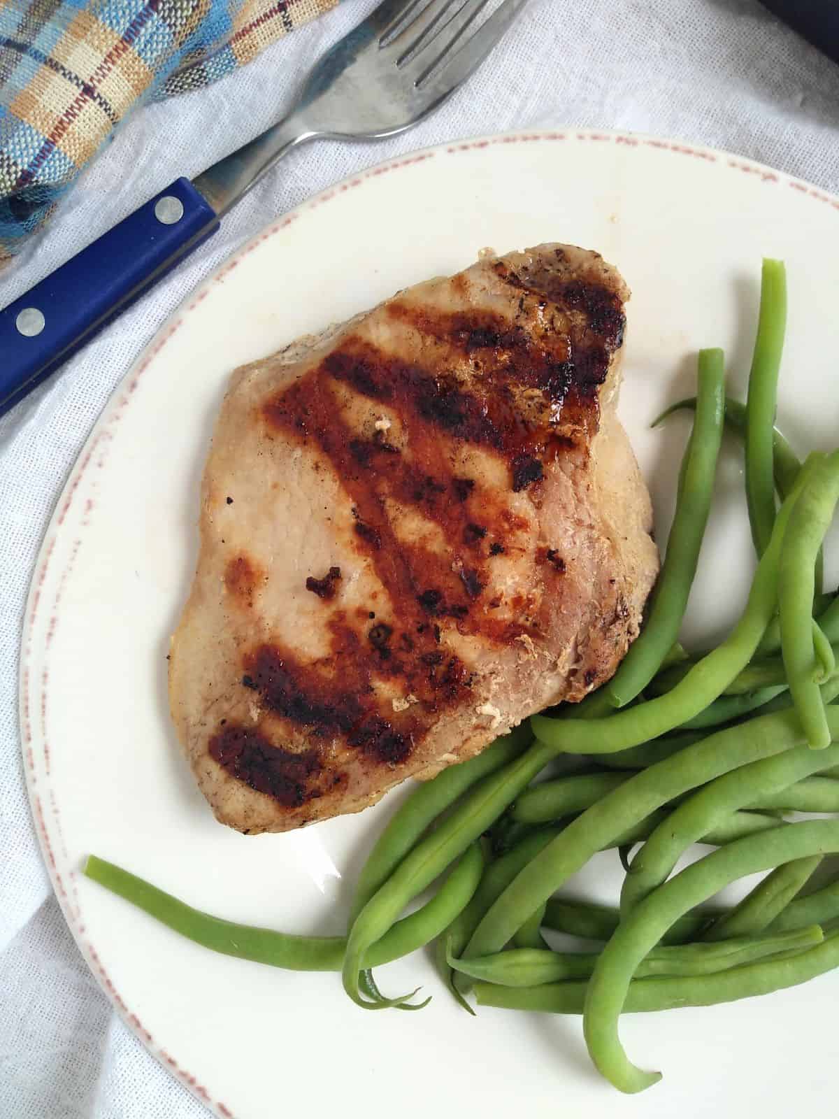 A grilled pork chop on a white plate with green beans. The plate is on a white towel with a blue/brown plaid towel on top and a blue handled fork