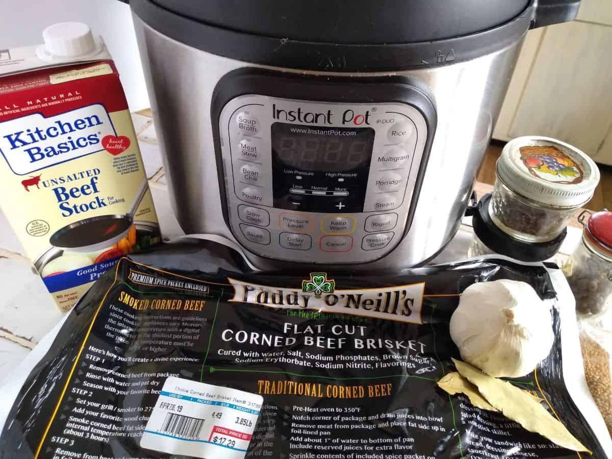 Ingredients for Instant Pot corned beef in front of the Instant Pot - Kitchen Basic Unsalted Beef Stock, Paddy O'Neill's Flat Cut Corned Beef Brisket, head of garlic, bay leaves, and jars of spices.