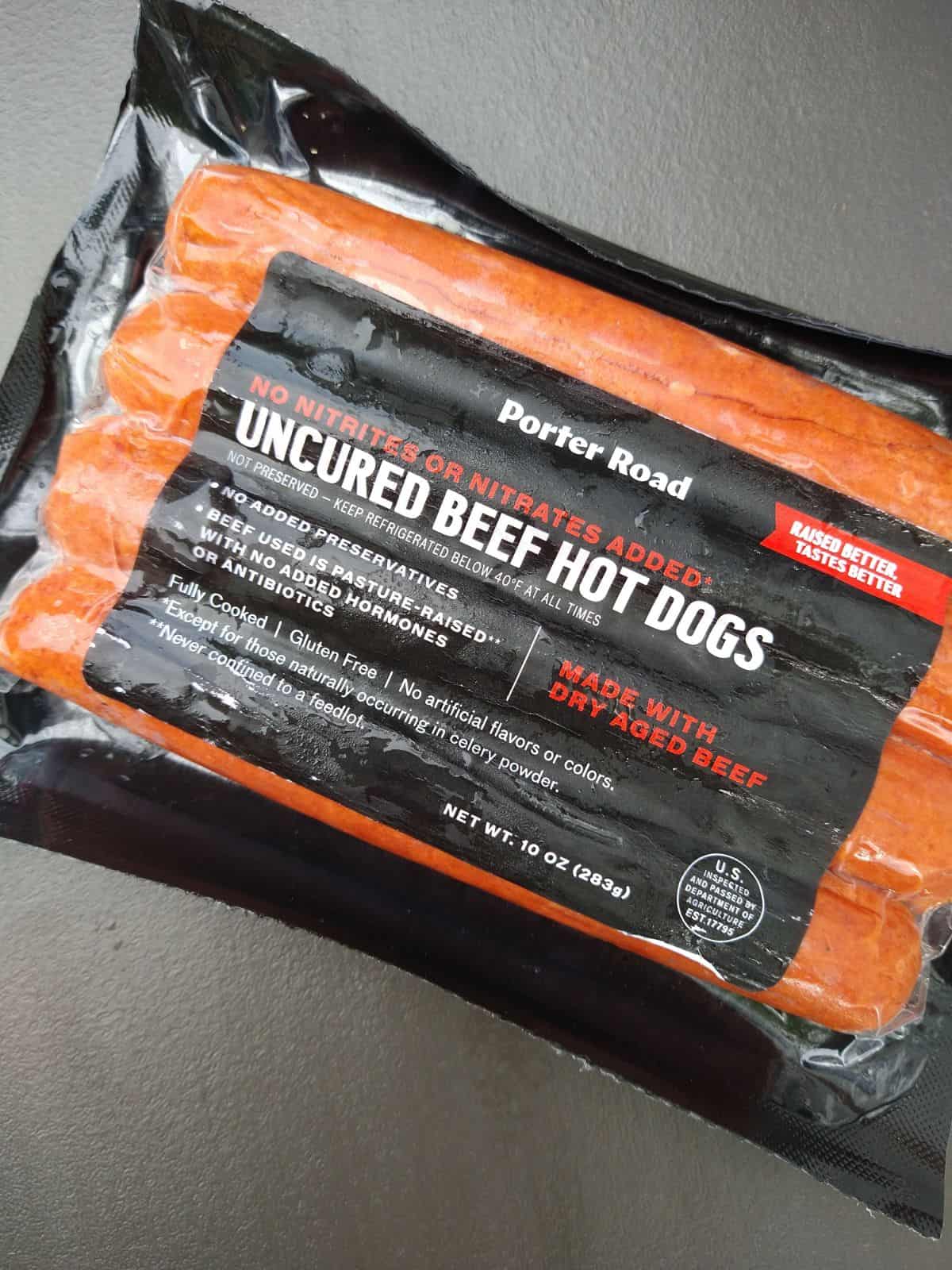 A package of Porter Road Uncured Beef Hot Dogs sitting on a gray colored table. There are 4 hot dogs in the package. 