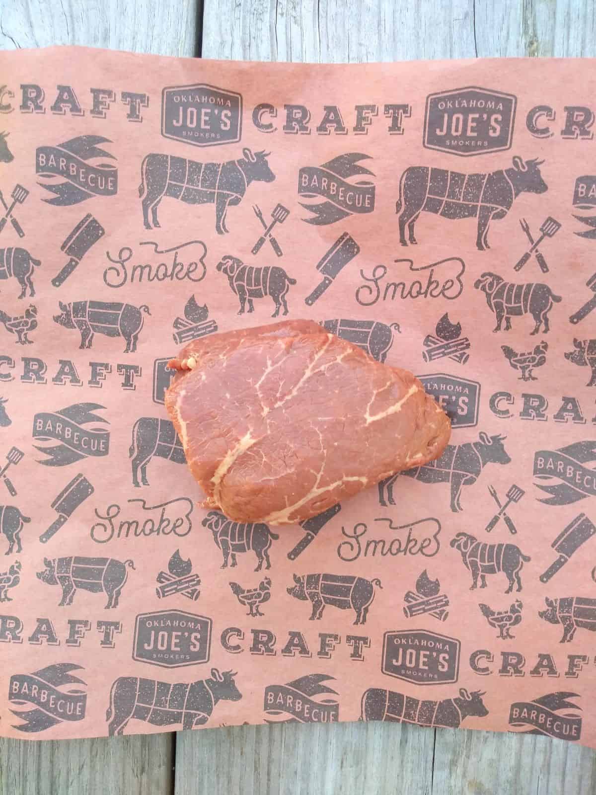 A raw top sirloin filet steak on a piece of buttering paper with grilling symbols on it, sitting on top of a wood picnic table.