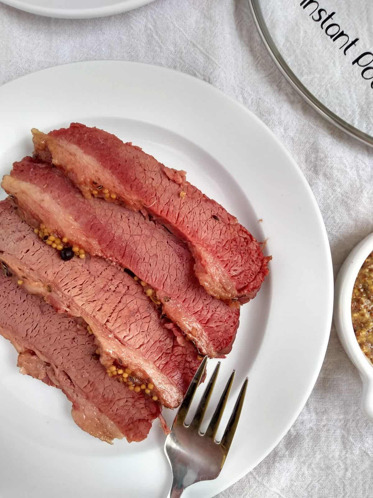 Sliced pieces of corned beef on a white plate on top of white towel with a glass Instant Pot lid in the top right corner. 