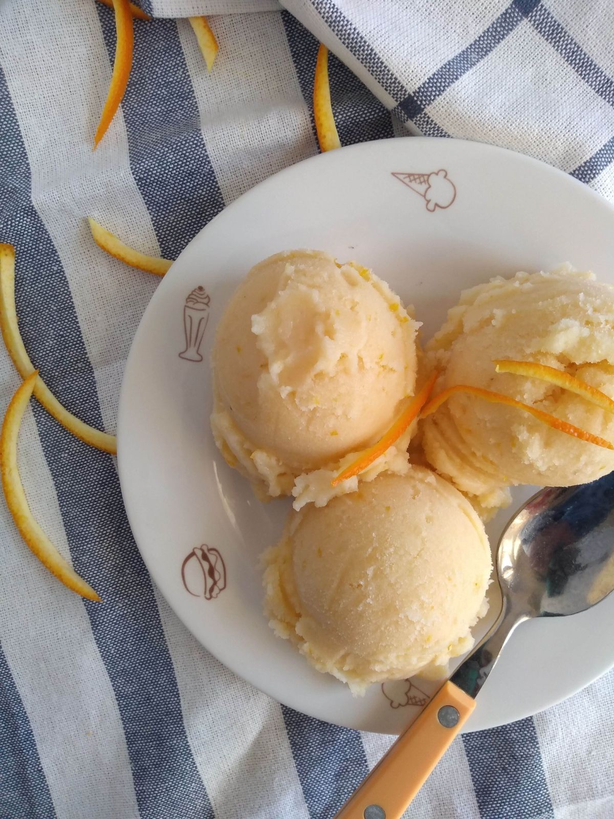 3 scoop of orange sherbet with orange zest on top, on a small white plate with pictures of ice cream sundaes and burgers along the edge. The plate is on a blue striped white towel.