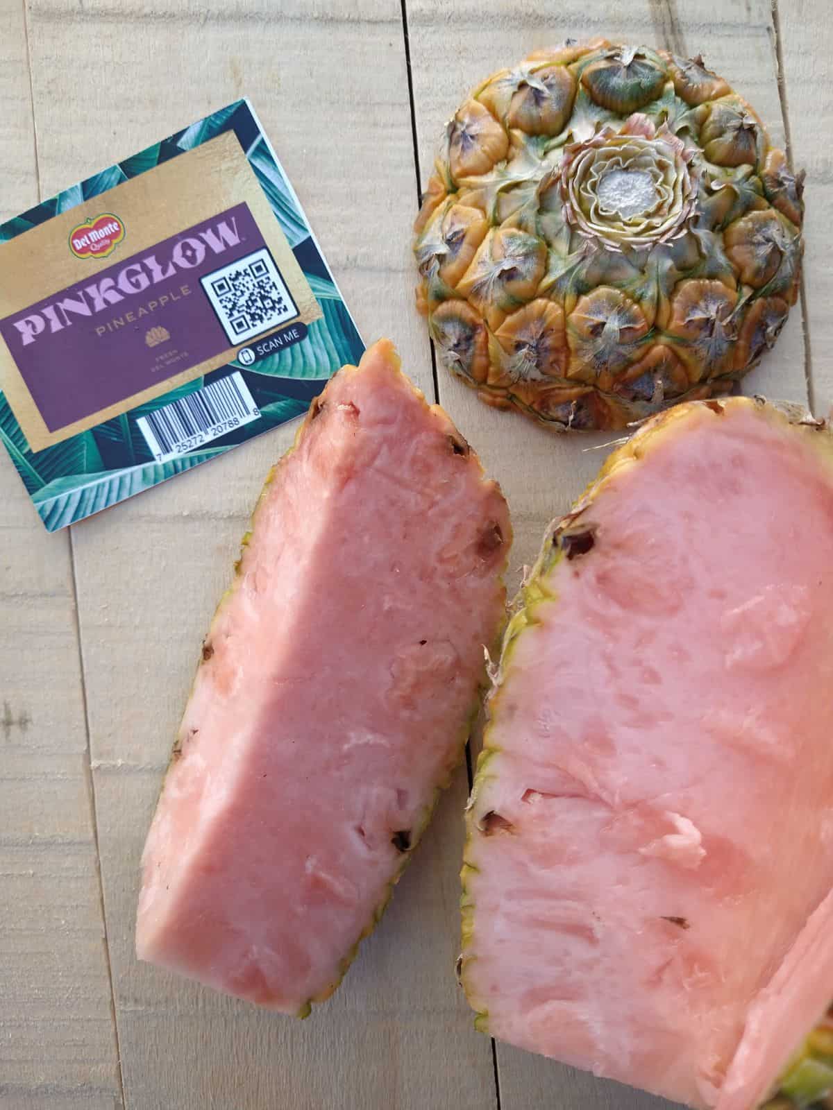 A wedge is cut out of a pink glow pineapple. The wedge and rest of pineapple are on a wood board with the top part of the pineapple - crown missing at the top. A tag is to the upper left corner.