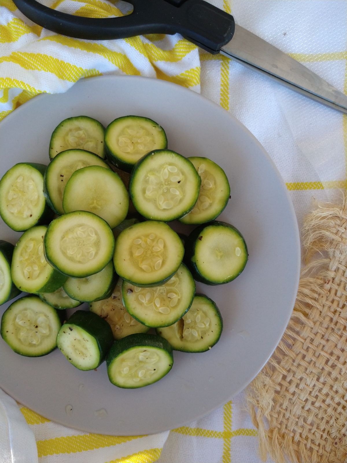 Slices of sous vide zucchini sitting on a gray plate on op of a white towel wit yellow strips and a piece of light brown burlap. There s a pair of scissors n the right top corner. 
