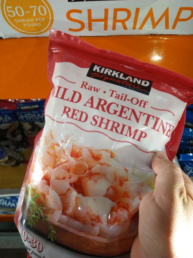 Holding a bag of Kirkland Raw Tail-Off  Wild Argentine Red shrimp in front of other bags of shrimp.